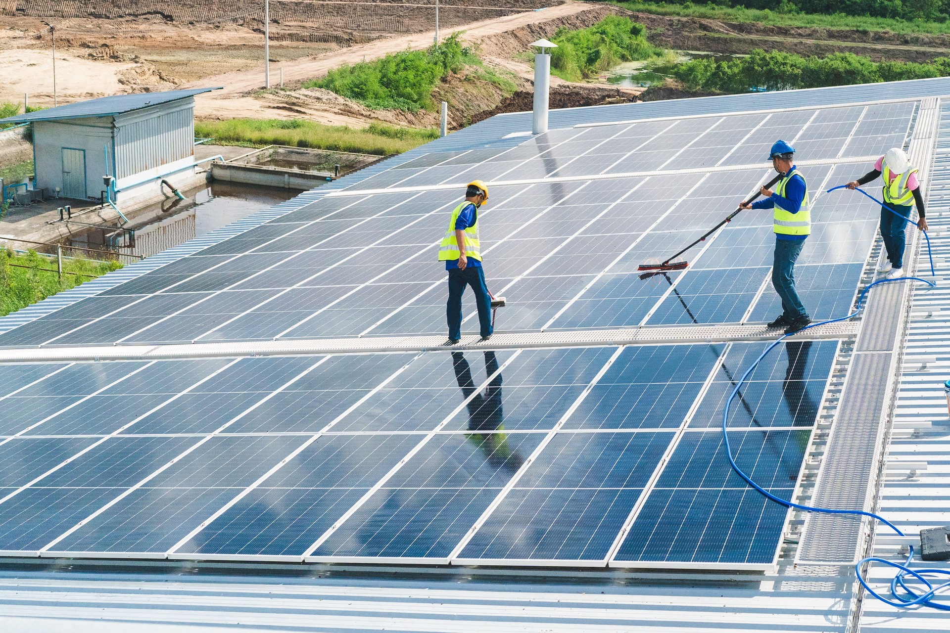 Professional worker cleaning solar panels with brush and washing with water on roof structure of building factory. Technician using mop to clean the dirty and dust, green electricity energy technology